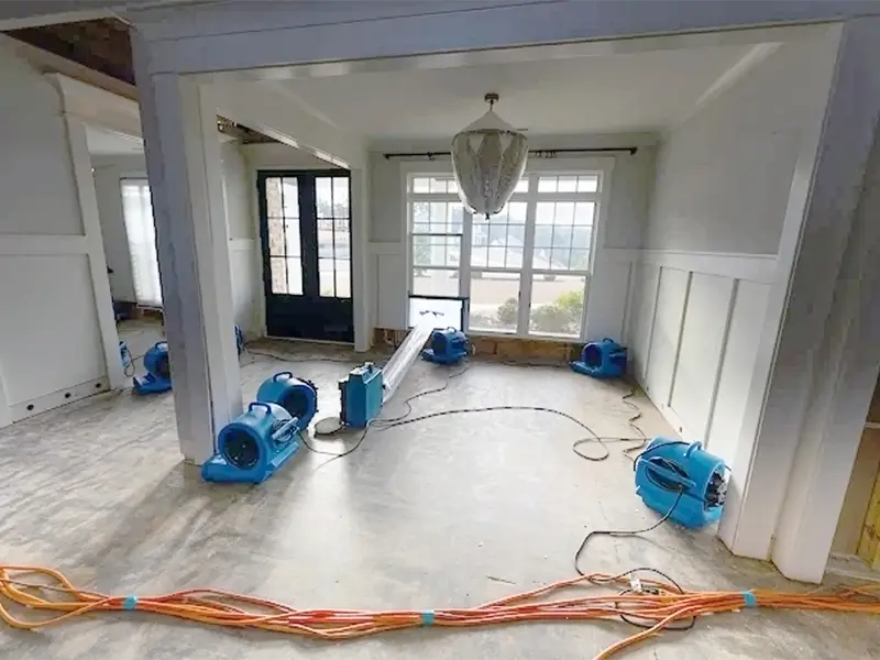 Blue air movers and a dehumidifier set up in a room with bare floors and white walls.