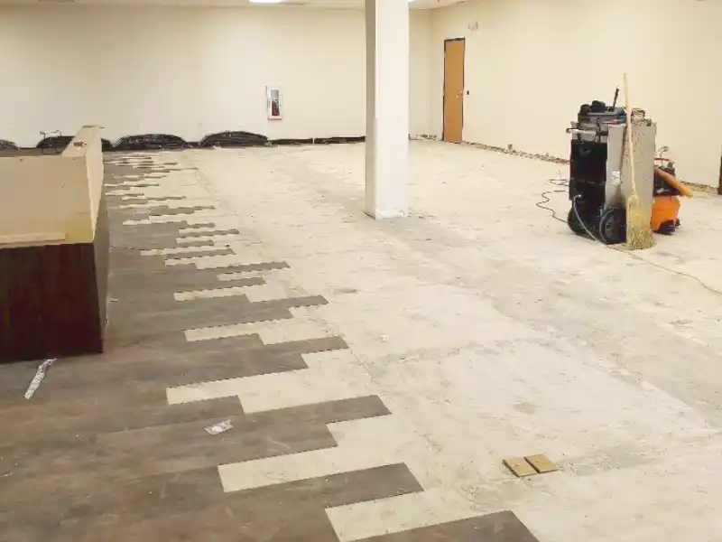 Partially installed wood flooring in a large empty room with tools and a broom nearby.
