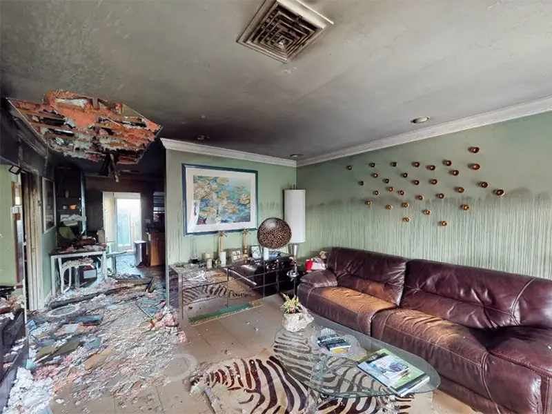 Living room with a damaged ceiling, debris on the floor, a brown leather couch, and a glass coffee table.