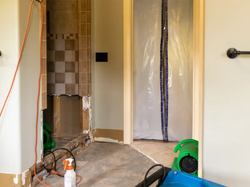 Bathroom under renovation with exposed wall, plastic doorway cover, and drying equipment.