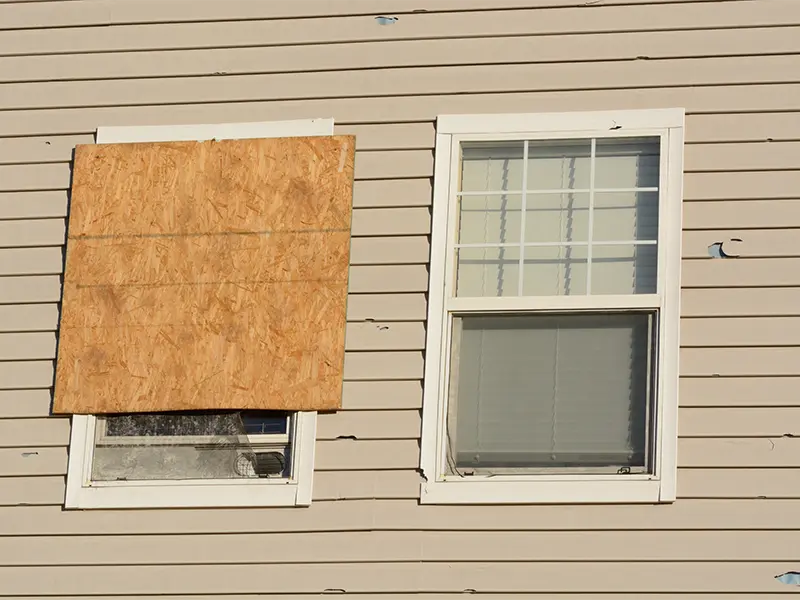 Two windows on a beige siding wall, one window partially covered with a plywood board.