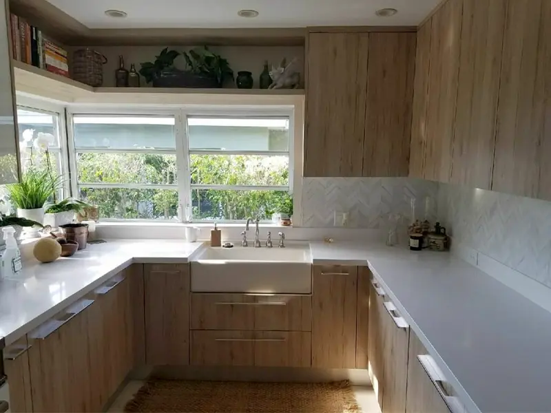 Modern kitchen with wooden cabinets, white countertops, a farmhouse sink, and large windows overlooking greenery.