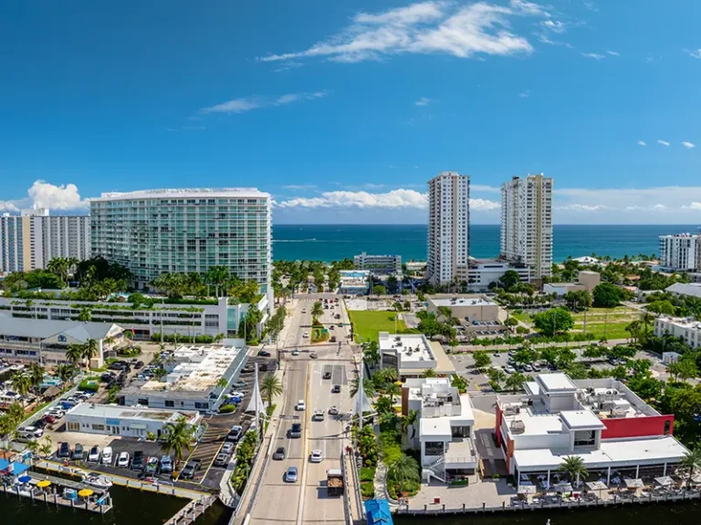 Coastal cityscape with high-rise buildings, a busy road, and the ocean in the background under a blue sky.