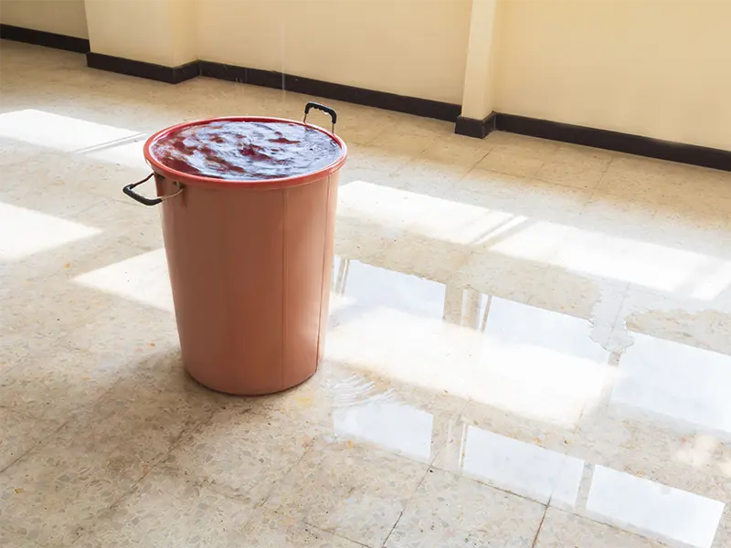 Large bucket overflowing with water on a tiled floor inside a room.